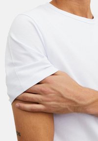 T-shirt en coton blanc avec des manches courtes, un col rond et des détails de couture visibles au niveau de l'ourlet des manches. Peau texturée et un tatouage sont partiellement visibles.