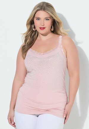 Frau mit langen blonden Haaren, die ein hellrosa ärmelloses Oberteil mit Strasssteinen und weiße Hosen trägt, stehend vor einem weißen Hintergrund.