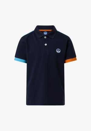 Garçon debout, les mains dans les poches, portant un polo bleu marine avec des manches à revers bleus et orange, un short cargo bleu marine, des chaussettes blanches et des baskets blanches.
