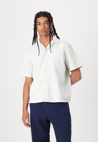 Chemise à manches courtes rayée dans des couleurs pastel légères, dotée d'un col pointu et d'une fermeture à boutons, portée avec un pantalon navy. Texture douce.