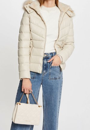 Vrouw draagt crème kleurige doorgestikte jas en witte top, blauwe jeans met hoge taille, houdt een crème doorgestikte handtas met gouden kettingbandjes vast.