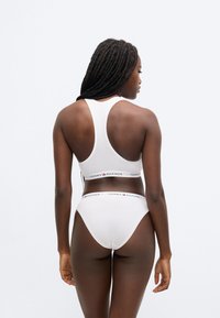 Valge puuvillane bralette ja tanga komplekt racerback disainiga, millel on logo ja elastne vöökoha riba. Sile tekstuur ja keha järgiv lõige.