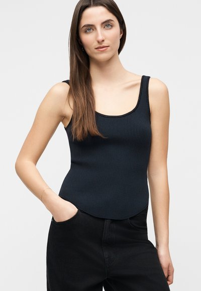 Νεαρή γυναίκα με μακριά καστανά μαλλιά, φοράει μαύρο ριμπ μπλουζάκι και μαύρο παντελόνι, έχοντας το ένα χέρι στην τσέπη, μπροστά σε απλό γκρι φόντο.