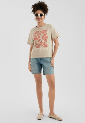 Beige short-sleeved t-shirt with red floral print and text "LES FLEURS," paired with denim shorts and woven beige slip-on shoes.