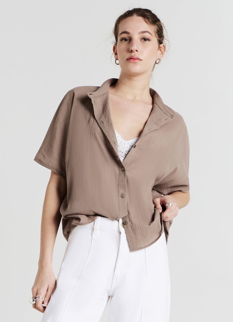 Chemise à manches courtes, boutonnée, en tissu texturé marron clair, avec une coupe décontractée et un col. Portée avec un pantalon blanc.