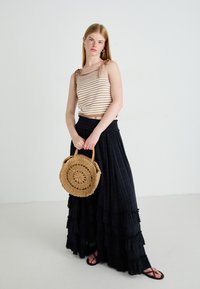 Femme jeune portant un haut sans manches à rayures et une longue jupe noire, tenant un sac à main en osier rond, se tenant devant un fond blanc uni.