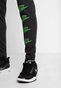 Graue Sporthosen mit neongrünen Logo-Akzenten an den Seiten, kombiniert mit schwarzen Sportschuhen, die ein silbernes Logo zeigen.