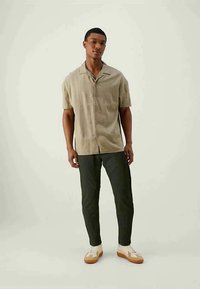 Jovem em pé, vestindo uma camisa de botão de mangas curtas bege, calças verde-escuras e ténis brancos com solas castanhas, contra um fundo claro e liso.