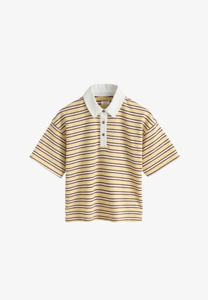 Dijete u žutoj prugastoj polo majici, svijetlim traper kratkim hlačama, crvenim čarapama i bež tenisicama stoji na bež pozadini.