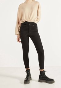 Jeans negros de tiro alto con cierre de cinco botones y bolsillos frontales, combinados con un suéter de punto texturizado beige y botas de combate negras con cordones.