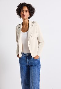 Hellbeige Blazer mit taillierter Passform, zwei vorderen Taschen und Revers, kombiniert mit lockeren blauen Denim-Jeans.