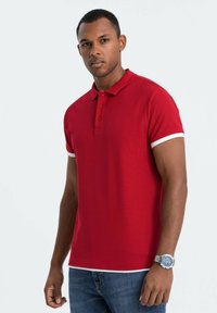 Polo rosso realizzato in tessuto testurizzato, con colletto, tre bottoni e dettagli bianchi sulle maniche. Abbinato a jeans blu.