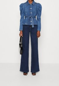 Džínová bunda s nařasenými rukávy, zapínáním na knoflíky a páskem v pase. Spárovaná se širokými džínovými kalhotami a držící černou kabelku.