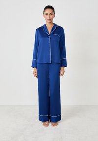 Ensemble de pyjama en satin bleu comprenant une chemise à boutons à manches longues, une poche poitrine et un pantalon large décoré de détails en passepoil blanc.
