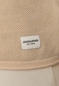 Beżowa dziana tkanina o tkanej fakturze, z prostokątną białą metką, na której znajduje się napis "JACK & JONES EST.1990" w czarnym kolorze w dolnej części.