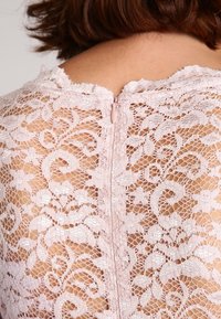 Ljust rosa spetsblus med intrikata blommönster, transparenta partier och en central dragkedja på baksidan för stängning.