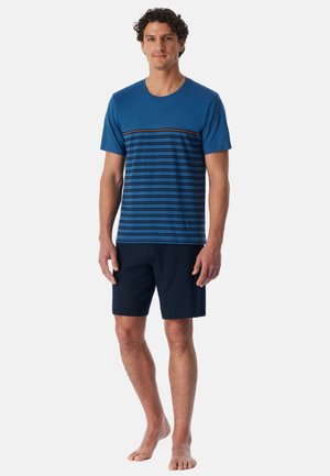 Mann steht barfuß, trägt ein blau gestreiftes Kurzarm-T-Shirt und dunkelblaue Shorts vor einem einfarbigen Hintergrund.