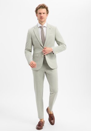 Homme marchant vers l'avant portant un costume gris clair, une chemise blanche, une cravate beige et des mocassins marron à glands sur fond blanc.