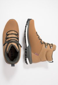 Bottes isolantes marron en tissu texturé, avec des lacets noirs et des semelles en caoutchouc. Équipées d'une boucle pour un enfilage facile et d'un col rembourré.