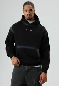 Zwarte hoodie van zachte stof, met een zak aan de voorkant, geribbelde manchetten en een klein roze logo op de borst.
