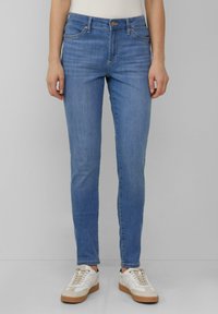 Jean en denim bleu clair, coupe slim, avec cinq poches et un délavage subtil, associés à des baskets blanches et beiges.