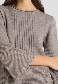 Brun stickad tröja med ribbad textur på bodice och breda, texturerade ärmar. Tyget ser mjukt och varmt ut.