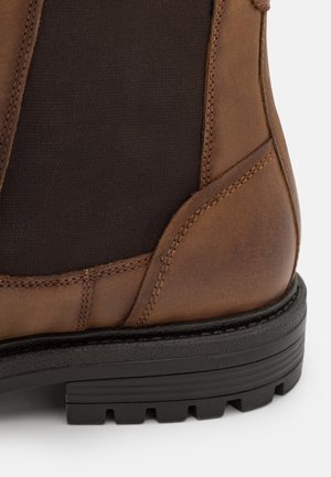 Gros plan sur le talon d'une botte en cuir marron avec un panneau latéral élastique noir et une semelle en caoutchouc noir épaisse à crampons profonds.