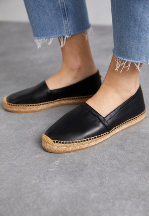 Füße, die schwarze Leder-Slip-on-Espadrilles mit Jutesohlen und blaue, ausgefranste Jeans auf grauem Boden tragen.