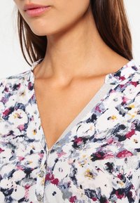 Chemisier fleuri avec un tissu léger, col en V, bordure argentée et fermeture par boutons, dans un mélange de couleurs rose, blanc et gris.