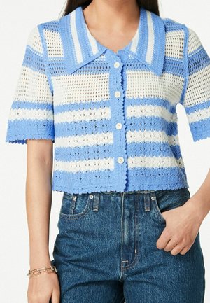 Vrouw draagt een kortarmig, blauw-wit gehaakt topje met knopen en een brede kraag, gecombineerd met high-waisted blauwe spijkerbroek.