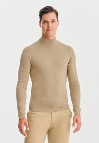 Beżowy dopasowany sweter z wysokim kołnierzem, wykonany z miękkiego materiału, z długimi rękawami i prążkowanymi mankietami, zestawiony z jasnobeżowymi, dopasowanymi spodniami. 