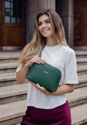 Zelená texturovaná kabelka se zlatým logem, se zipovým uzávěrem a nastavitelným popruhem. Model má na sobě bílý top a vínovou sukni.