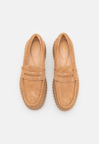 Mocasines de ante en color beige claro con punta redonda, que presentan un detalle de costura y suela de goma con un diseño texturizado. El interior incluye perforaciones.
