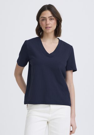 Frau trägt ein navyblaues T-Shirt mit V-Ausschnitt und kurzen Ärmeln sowie weiße Hose, steht mit einer Hand hinter dem Rücken vor einem schlichten Hintergrund.