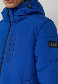 Niebieska kurtka puchowa z zamkiem błyskawicznym, regulowanym kapturem i małą kieszenią na piersi. Na rękawie znajduje się markowa naszywka. Gładka tekstura.