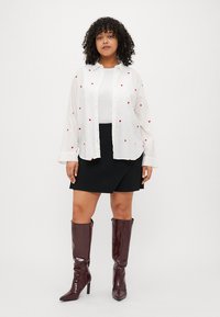 Λευκό πουκάμισο με κομπιά ζαχαρωμένα σχέδια, μαύρη σατέν φούστα, και γυαλιστερά μποτάκια σε μπορντό απόχρωση μέχρι το γόνατο. Λείες υφές και άνετη εφαρμογή.