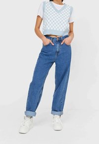 Chaleco de punto a cuadros azul y blanco, cortado, sobre una camiseta blanca, combinado con unos jeans de mezclilla azul de cintura alta. Mangas enrolladas y zapatillas blancas.