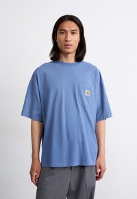 Blå bomulds T-shirt med rund hals, korte ærmer og en brystlomme med et logo label. Stoffet ser blødt og utextureret ud.