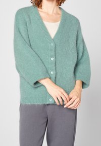Cardigan en peluche teal avec un col en V, des manches oversized et six boutons bleu clair. Porté par-dessus un haut beige clair, associé à un pantalon gris.