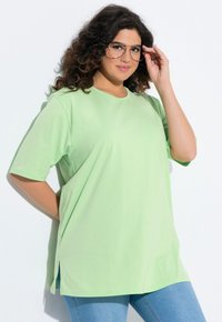 Hellgrünes, oversized T-Shirt aus weicher Baumwolle mit kurzen Ärmeln, rundem Ausschnitt und seitlichen Schlitzen. Kombiniert mit blauen Jeans.