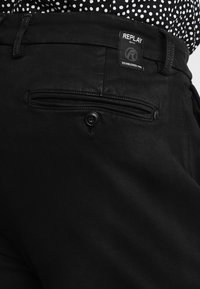Pantalones de mezclilla negros con un bolsillo trasero con cremallera, que presentan una etiqueta con el nombre "REPLAY." Textura suave y corte ajustado.