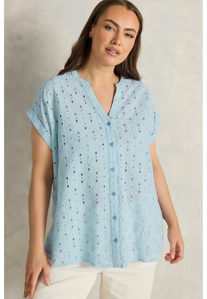 Donna che indossa una blusa azzurro chiaro a maniche corte con bottoni e motivi floreali a occhiello, abbinata a pantaloni bianchi, su uno sfondo neutro.
