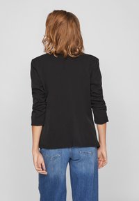 Blazer negro con mangas tres cuartos arrugadas, textura suave y abertura en la espalda, combinado con pantalones vaqueros de pierna ancha azules.