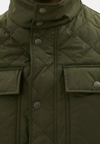 Veste verte matelassée avec un col haut, des boutons-pression et deux poches avant. Présente une texture lisse et un motif de coutures diagonales.