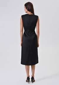 Donna in piedi di spalle, che indossa un vestito midi nero senza maniche con zip sul retro e scarpe con tacco alto nere su uno sfondo semplice.