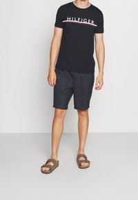 Muž v černém tričku Tommy Hilfiger, tmavých kostkovaných šortkách a hnědých sandálech stojí na jednobarevném bílém pozadí.