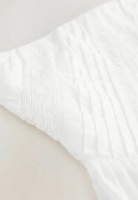 Bílá látka s texturovanými vertikálními liniemi a jemným krajkovým zdobením podél jednoho okraje. Měkký a lehký materiál.