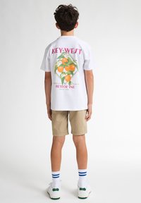 Witte t-shirt met korte mouwen met daarop een kleurrijke oranje graphic en de tekst "KEY WEST" op de achterkant, gecombineerd met beige shorts en witte sneakers.