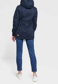 Navy insulated jacket with a hood, drawstring waist, and side accents. Worn over denim jeans and white sneakers, showing a sporty look.