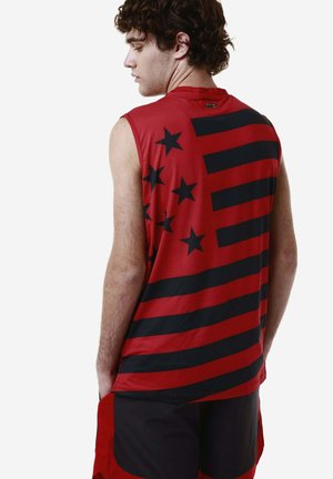 Jeune homme aux cheveux bouclés portant un haut rouge sans manches orné d'étoiles et de rayures noires, debout les mains dans les poches, de dos.